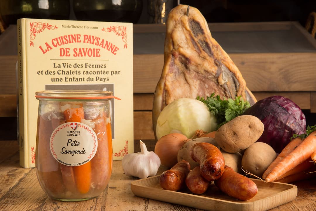 La potée savoyarde – Une recette locale emblématique  Voici l’une de nos recettes phares : la potée savoyarde, un plat mijoté généreux et réconfortant, fidèle à la tradition culinaire de nos montagnes.  En arrière-plan, vous apercevez l’un des 23 exemplaires originaux du livre La Cuisine paysanne de Savoie de Marie-Thérèse Hermann, véritable trésor d’archives dont nous nous inspirons pour faire revivre les saveurs d’antan.  Sur la droite de l’image, sont mis en valeur les ingrédients clés de cette préparation : des légumes de saison, les fameux diots savoyards, ainsi qu’un morceau de viande séchée suspendu, comme dans les fermes d'autrefois.  L’atmosphère qui se dégage de cette scène est résolument rustique, authentique… et résolument gourmande.